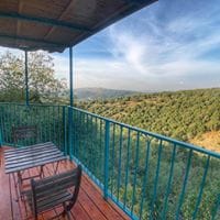 A private balcony overlooks lush green hills, framed by a blue railing. Two black chairs and a small table are positioned on the wooden deck, creating an inviting space for enjoying the expansive views of the surrounding forest and landscape.