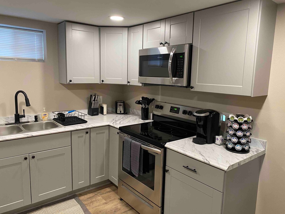 A well-equipped kitchen features modern gray cabinetry and a marble countertop. Stainless steel appliances, including a microwave and oven, are visible alongside a coffee maker and a selection of coffee pods. A double sink and drying rack are included for convenience.