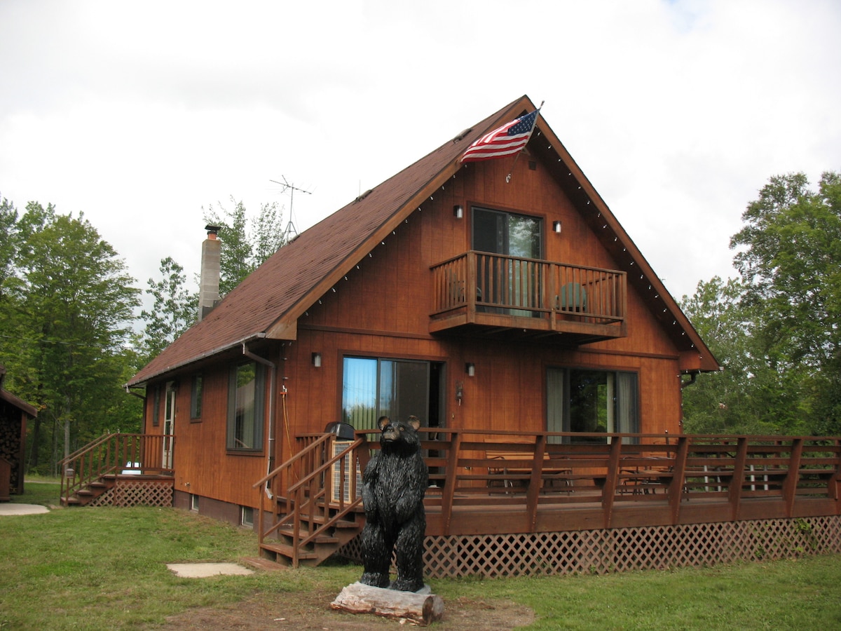 The chalet's exterior features a warm wooden façade punctuated by a triangular roof. A balcony extends from the upper level, with an American flag displayed prominently. A wooden bear sculpture stands at the forefront, adding a rustic touch to the surroundings of green grass and trees.