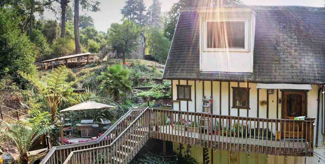 A charming two-story homestead is surrounded by lush greenery and towering trees. A wooden deck wraps around the ground floor, featuring an outdoor seating area underneath a large umbrella. Natural light enhances the inviting atmosphere of the home.