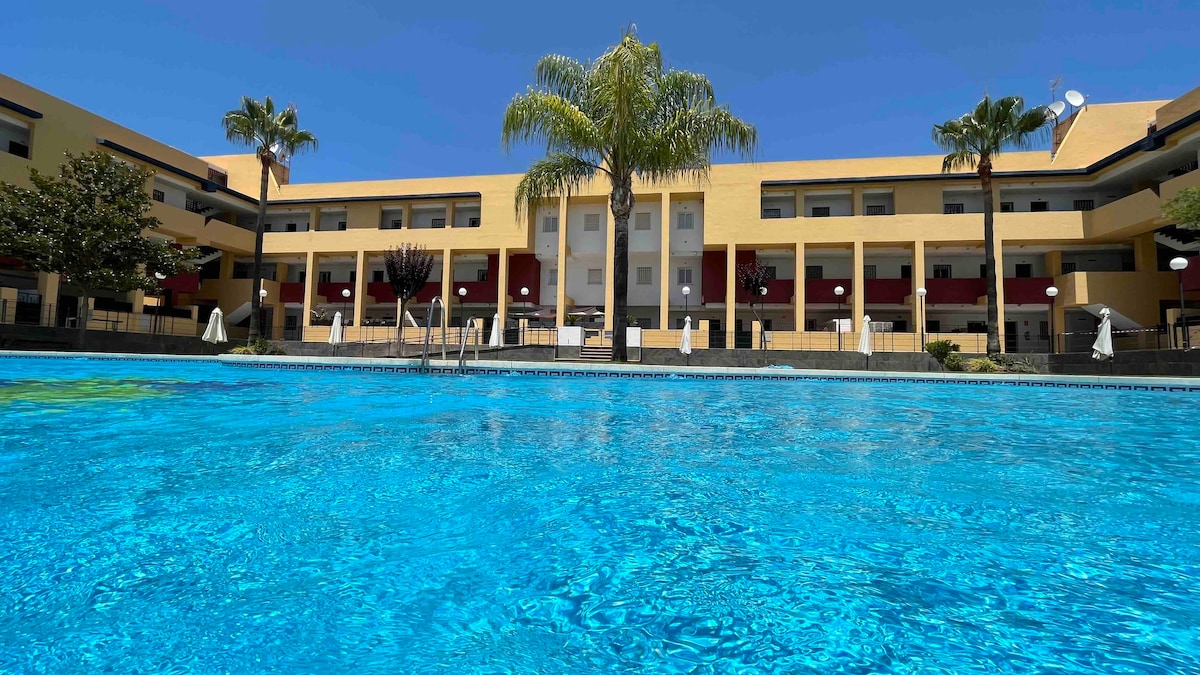 A tranquil salt-water pool reflects a vibrant blue, with tall palm trees framing the inviting outdoor space. A spacious building stands in the background, featuring multiple balconies and large windows. Sun loungers are arranged around the pool, offering a serene environment for relaxation.