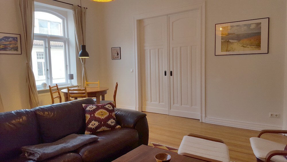 A living area features a comfortable dark leather sofa, complemented by a colorful patterned cushion. A round dining table with four wooden chairs is visible, alongside a stylish floor lamp. Two decorative wall frames add character, while natural light fills the space through a large window.