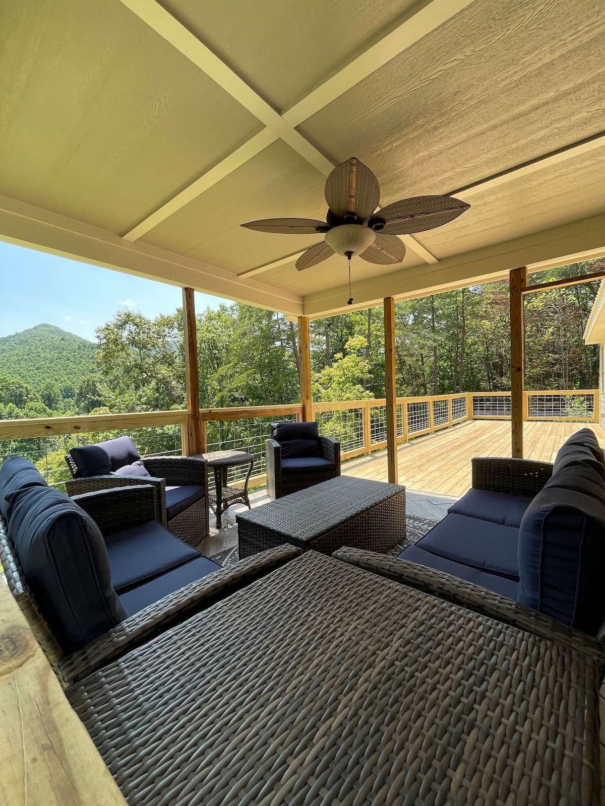 A spacious deck features comfortable wicker seating arranged around a coffee table. A ceiling fan is positioned overhead, enhancing airflow. Lush trees and distant mountains provide a serene backdrop, emphasizing the inviting outdoor living space.