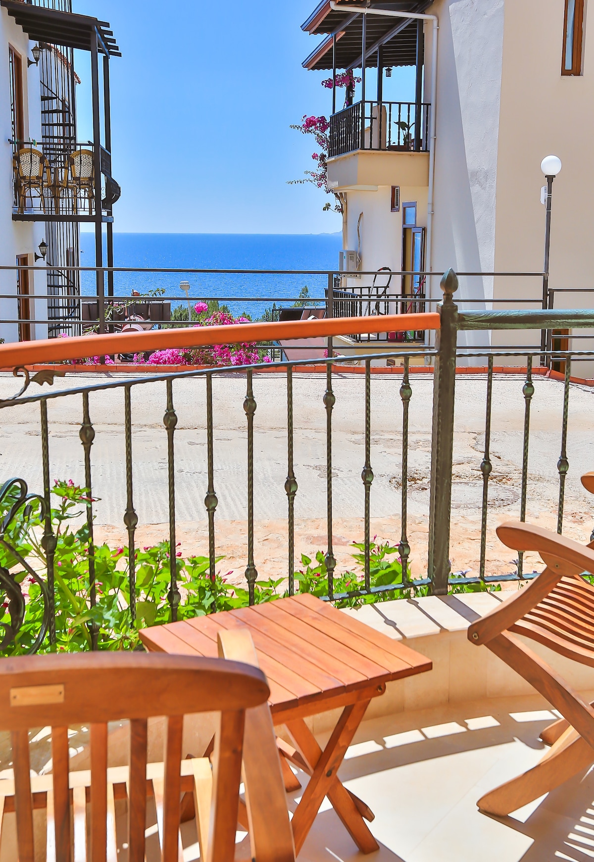 A balcony offers a view of the sea, framed by blooming flowers. Two wooden chairs and a small table are positioned on the balcony, providing a spot to relax. The surrounding buildings feature white walls and iron railings, enhancing the coastal ambiance.