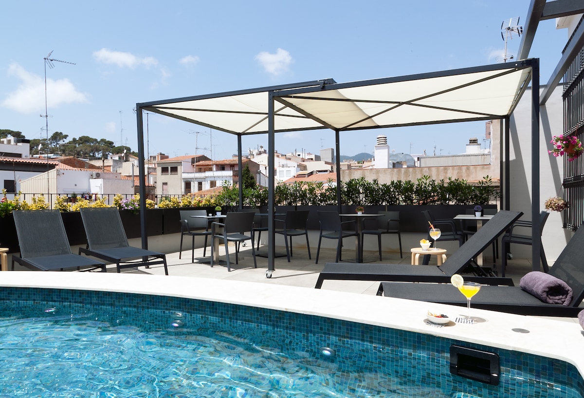 A rooftop terrace features a pool with a mosaic tiled edge, surrounded by sun loungers and a shaded seating area. Potted plants line the space, and tables with chairs are set up for dining or relaxation. Drinks are placed on small tables, complementing the inviting atmosphere.