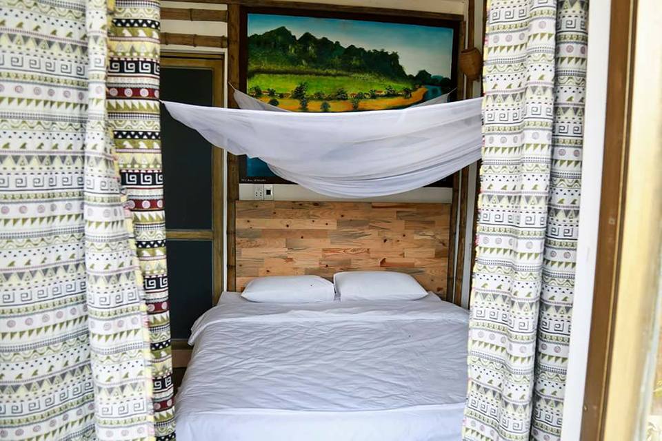 A cozy sleeping area features a spacious bed dressed in white linens. Soft mosquito nets are draped over the bed, providing a serene atmosphere. The environment is enhanced by patterned curtains and a large painting depicting a lush landscape.