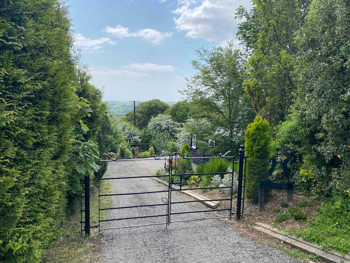 A gated entrance reveals a gravel driveway leading through lush greenery. Trees and shrubs frame the path, offering glimpses of the surrounding landscape beyond, while the sky above features soft clouds, contributing to a serene atmosphere.