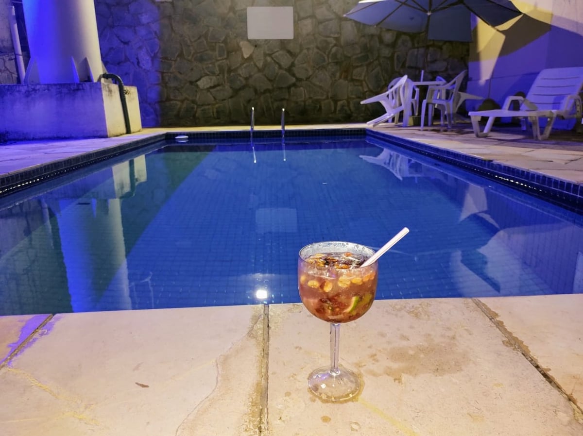 A refreshing drink is placed beside a shimmering pool, reflecting soft blue lights. Comfortable white lounge chairs are arranged nearby, providing a serene spot for relaxation. The surrounding area features textured stone walls, enhancing the tranquil ambiance of the outdoor space.
