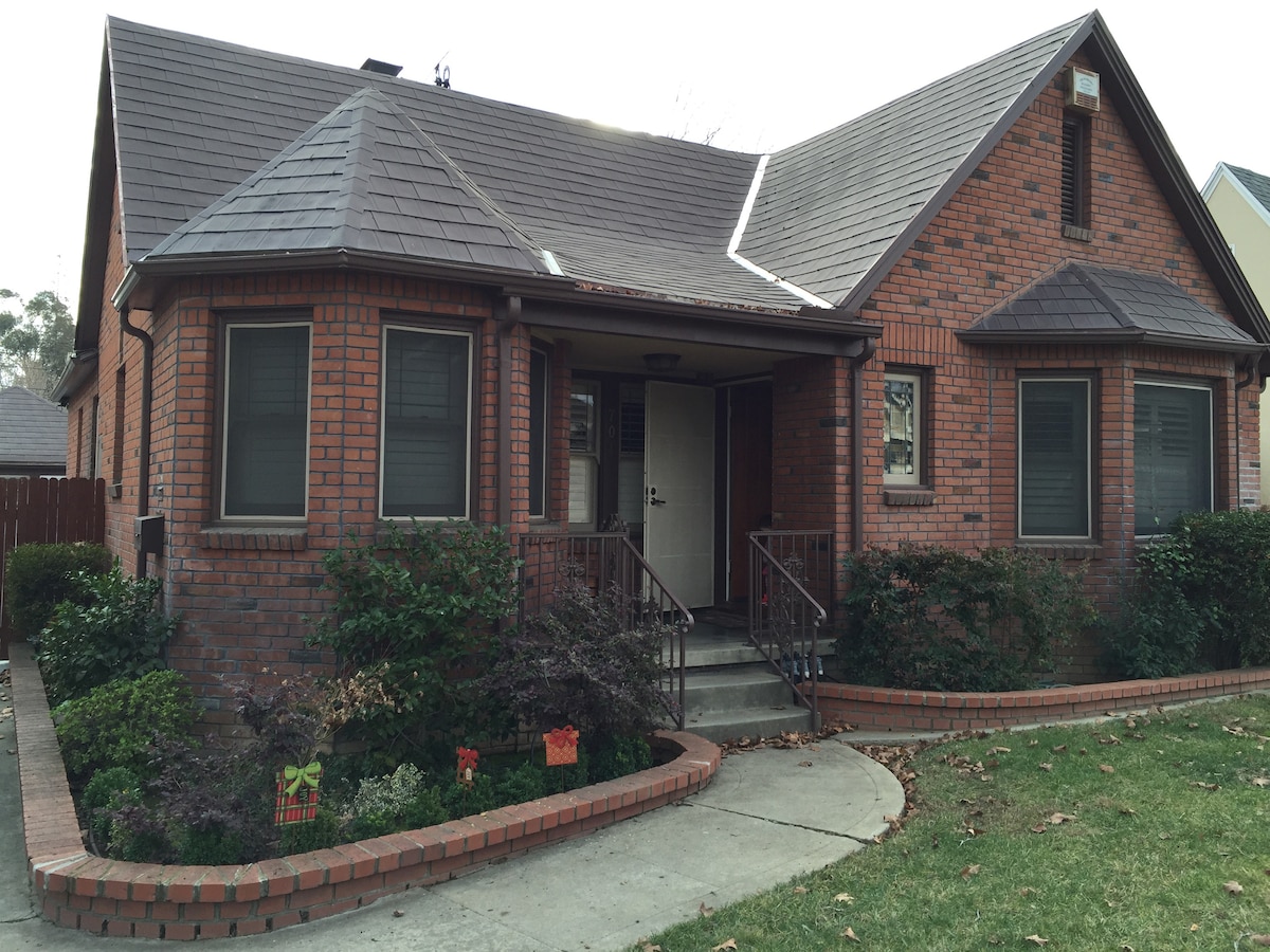 The exterior of a brick guest house is highlighted, showcasing a gently sloped roof and large front steps. Lush green plants and small decorative elements frame the entrance, while windows with grid patterns offer natural light. Soft landscaping leads to the front patio.