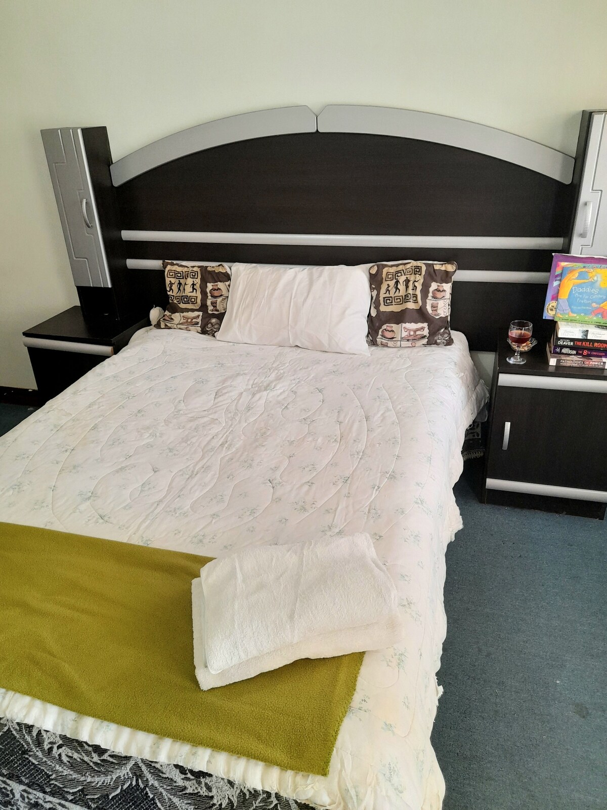 A comfortable sleeping area features a spacious bed with a light floral-patterned coverlet and decorative pillows. A green throw is neatly placed at the foot of the bed. A bedside cabinet holds a glass and a collection of books, providing a welcoming touch.
