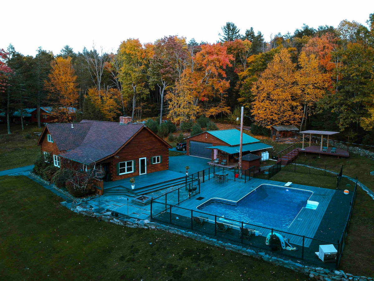 A spacious log cabin with a well-maintained outdoor area is surrounded by colorful autumn foliage. A fenced-in swimming pool is visible, along with various decks and pathways leading to additional structures, adding to the inviting outdoor setting.