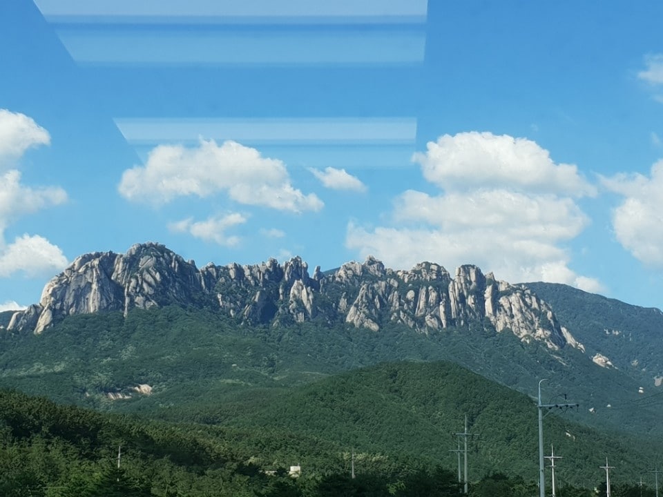 A striking view of Ulsan Rock is captured from a large window, with a backdrop of clear blue skies and scattered clouds. The rocky mountain range stands majestically against lush green hills, creating a picturesque natural landscape.