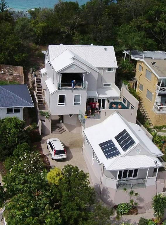 An aerial view of a dual-level cottage showcases a contemporary design with a white roof and solar panels. Surrounding greenery provides a natural setting, while the ocean can be seen in the background. A driveway leads to a parked vehicle adjacent to the entrance.