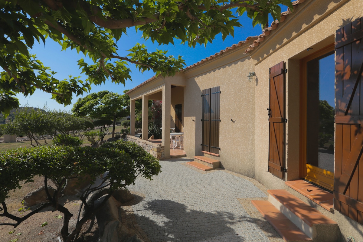 An exterior view of a residence is presented, featuring a stone pathway leading to the main entrance. Well-maintained greenery surrounds the property, and wooden shutters adorn the windows. Sunlight enhances the warm tones of the building's façade and highlights the inviting atmosphere.