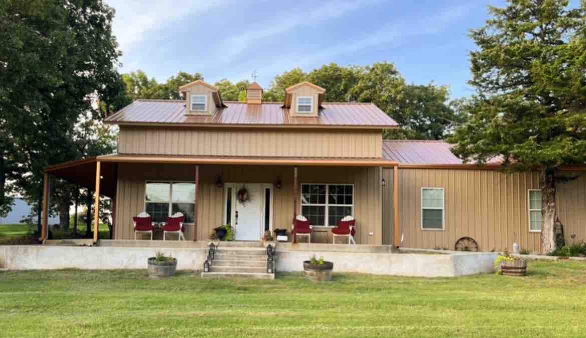 A welcoming exterior showcases a two-story house with a metal roof and a covered porch. The porch is furnished with several red chairs and potted plants, framed by green lawn and trees, creating an inviting outdoor space.