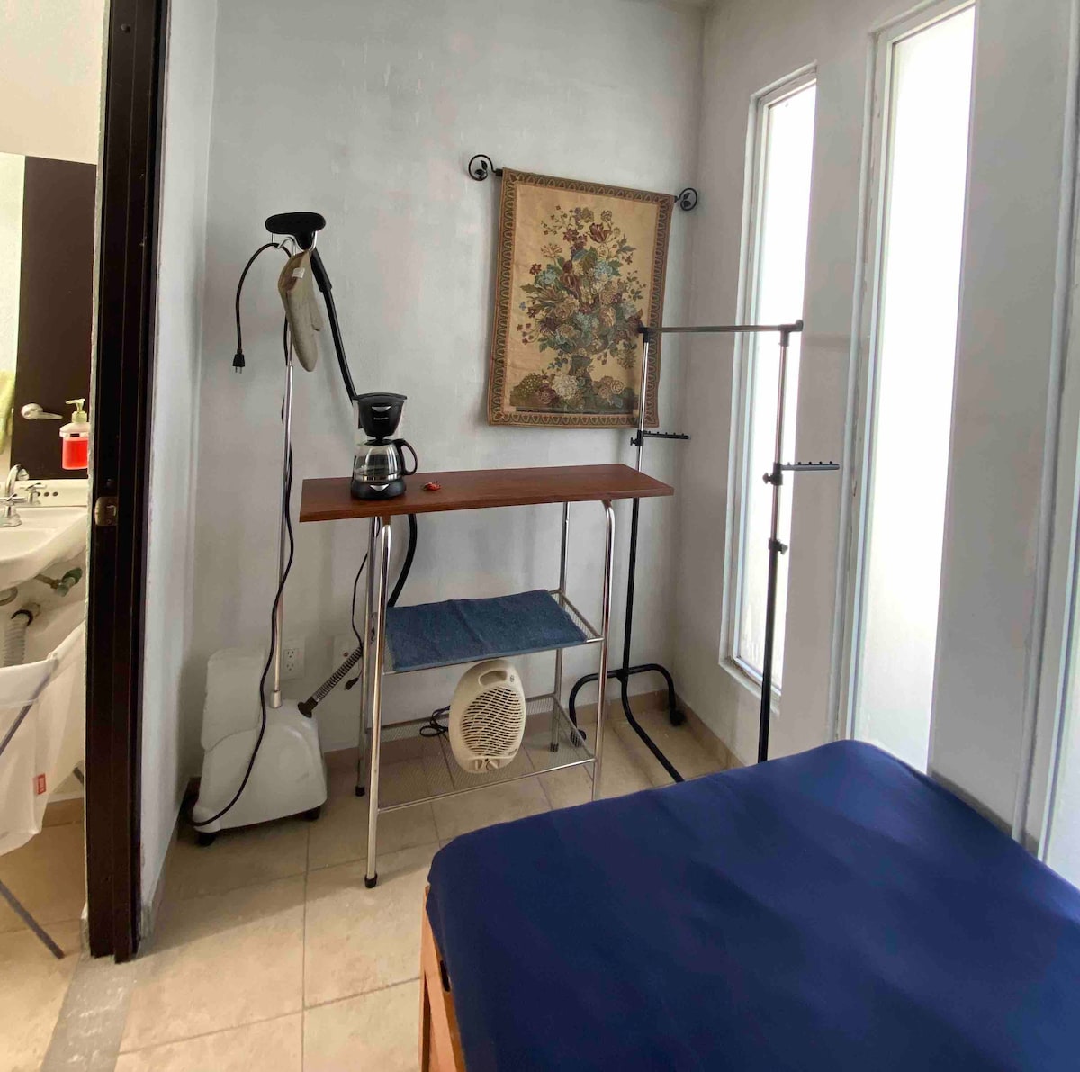 A cozy room features a spacious bed with a blue coverlet. A small wooden table accommodates a coffee maker, while a clothing rack provides hanging space. Natural light streams through three windows, enhancing the fresh ambiance. A bathroom area is visible in the background.