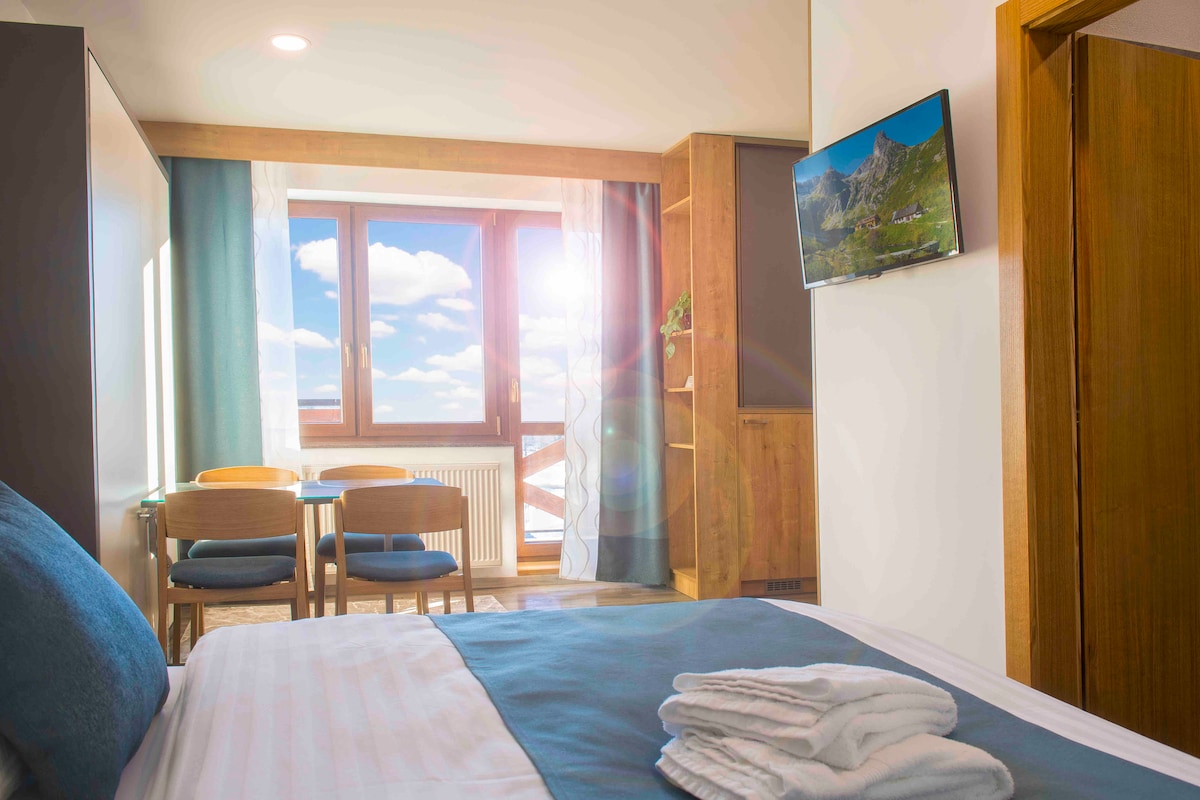 A bright sleeping area features a spacious bed with folded towels neatly arranged on top. A table with two chairs is positioned near a large window, allowing natural light to fill the room. A television is mounted on the wall, offering convenient entertainment.