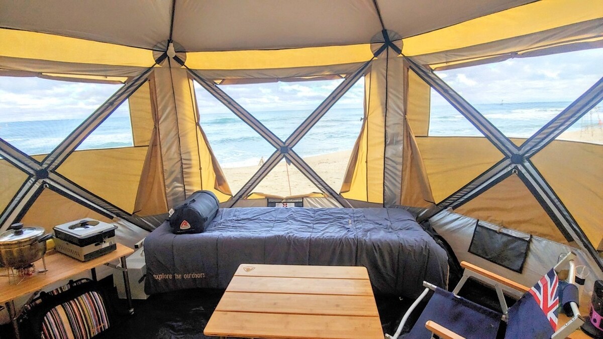 The interior of a spacious tent features large windows that provide views of the beach and ocean. A comfortable bed is set against one wall, accompanied by a wooden table and seating area. Natural light fills the space, enhancing the camping experience.