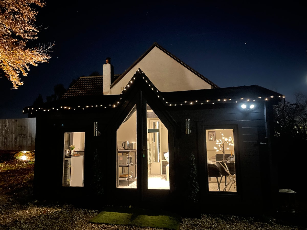 The exterior of a modern cabin is illuminated at night, with soft lighting accentuating its architecture. Large glass doors provide views into the living space, while decorative lights adorn the roofline, creating a welcoming ambiance. Surrounding trees and the night sky add to the serene atmosphere.