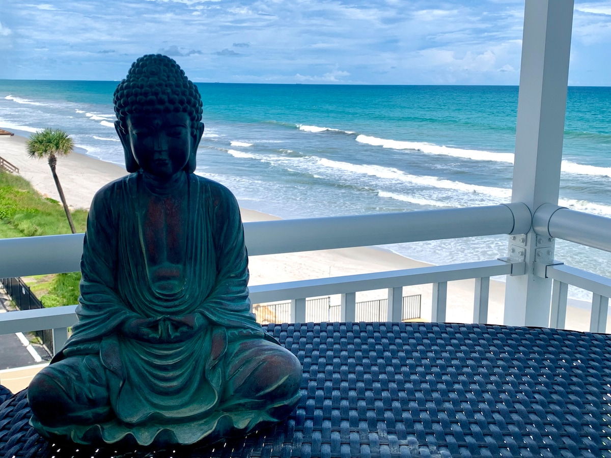 A serene Buddha statue is positioned on a dark woven table, overlooking the ocean. The ocean waves gently lap at the sandy shore under a cloudy sky, with palm trees visible in the distance. The scene captures a peaceful coastal atmosphere.
