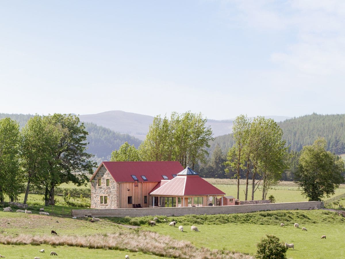 A modern cottage showcasing a blend of stone and wood construction is set against a backdrop of gentle hills. The bright red roof complements the green landscape, while large windows invite natural light and offer panoramic views of the serene surroundings.