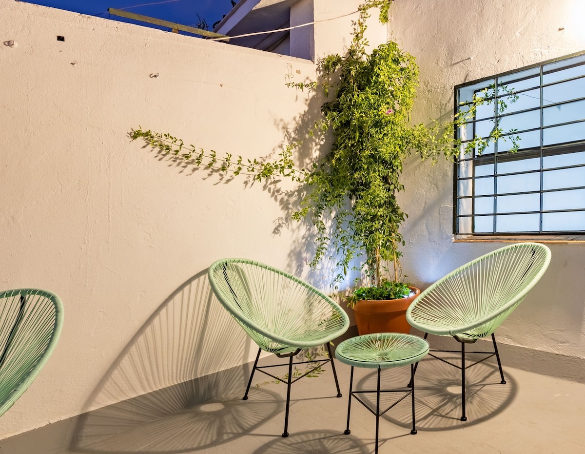 A serene terrace features two light green woven chairs and a matching small table set against a white wall. A potted plant adds natural beauty, casting delicate shadows on the ground. Soft lighting enhances the tranquil ambiance of the outdoor space.