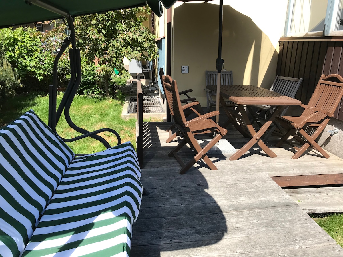 A stylish outdoor area features a striped swing and a wooden dining table surrounded by several folding chairs. The space is complemented by a well-maintained lawn and greenery in the background, offering a serene setting for relaxation.