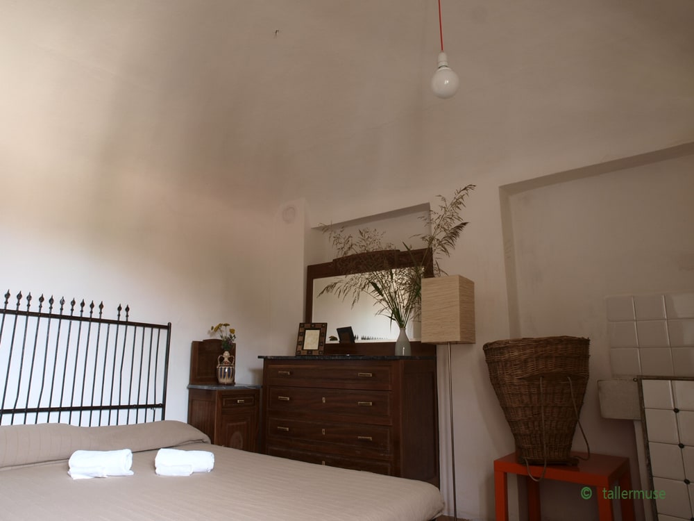 A spacious room features a simple double bed with white towels neatly arranged on top. A wooden dresser and a woven basket add to the decor. A mirror reflects subtle natural elements, while a single pendant light hangs from the ceiling, enhancing the airy feel of the space.