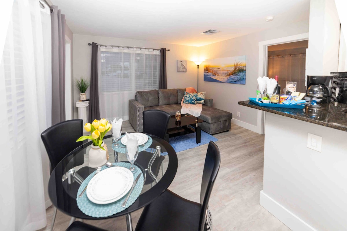 An open-concept living area features a modern grey sofa paired with a decorative throw and pillows. A glass dining table is set for four with a blue placemat and white dishes, while natural light filters through sheer curtains. A small decorative plant and wall art enhance the space.