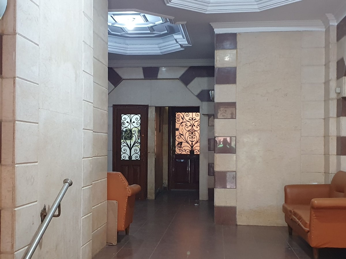 The entrance hall features a sleek design with polished stone walls and a stylish ceiling. Two wooden doors with intricate ironwork are visible, leading to separate areas. Comfortable brown sofas are placed along one side, offering a welcoming area for guests.