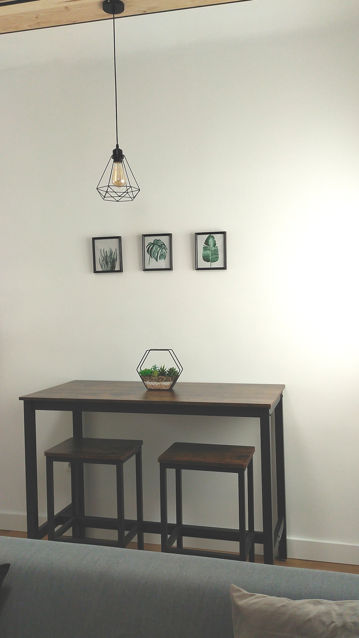 A small dining area presents a tall wooden table paired with two stools. Above the table, a geometric pendant light casts a soft glow. Three framed botanical prints are arranged on the wall, adding a touch of greenery to the space.