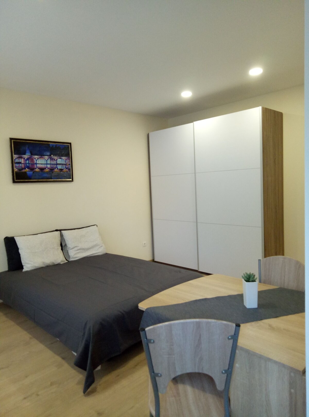 A cozy sleeping area is present, featuring a spacious bed with a dark blanket and decorative cushions. A wooden table and chairs provide a dining space, while a compact wardrobe stands nearby. Soft lighting enhances the room's inviting atmosphere.