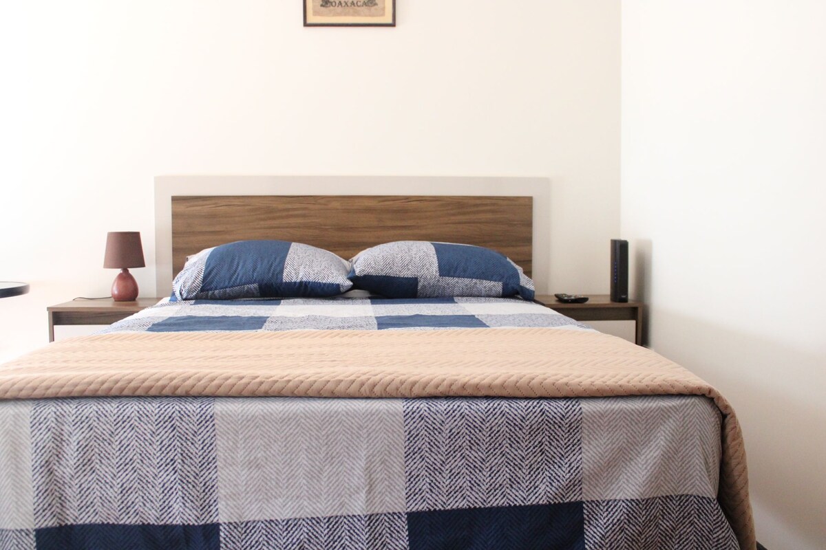 A comfortable sleeping area is presented, featuring a double bed with a patterned blanket. Nightstands with lamps flank the bed. A sleek piece of furniture holds a remote control and a speaker, while minimalistic decor maintains a clean aesthetic.