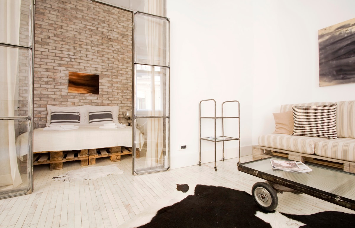 A spacious bedroom area is separated by glass partitions, featuring a cozy bed with a neutral-colored duvet and pillows. A modern sofa with striped upholstery complements the space. A rustic wooden cart serves as a coffee table, while a decorative cowhide rug adds texture to the floor.