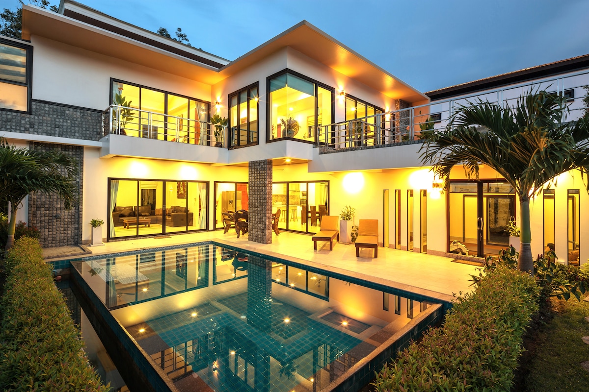 The villa's exterior is highlighted by a spacious terrace overlooking a private pool. Large glass doors create seamless connectivity between indoor and outdoor living areas. Lush greenery surrounds the property, while warm lighting creates an inviting ambiance during twilight.
