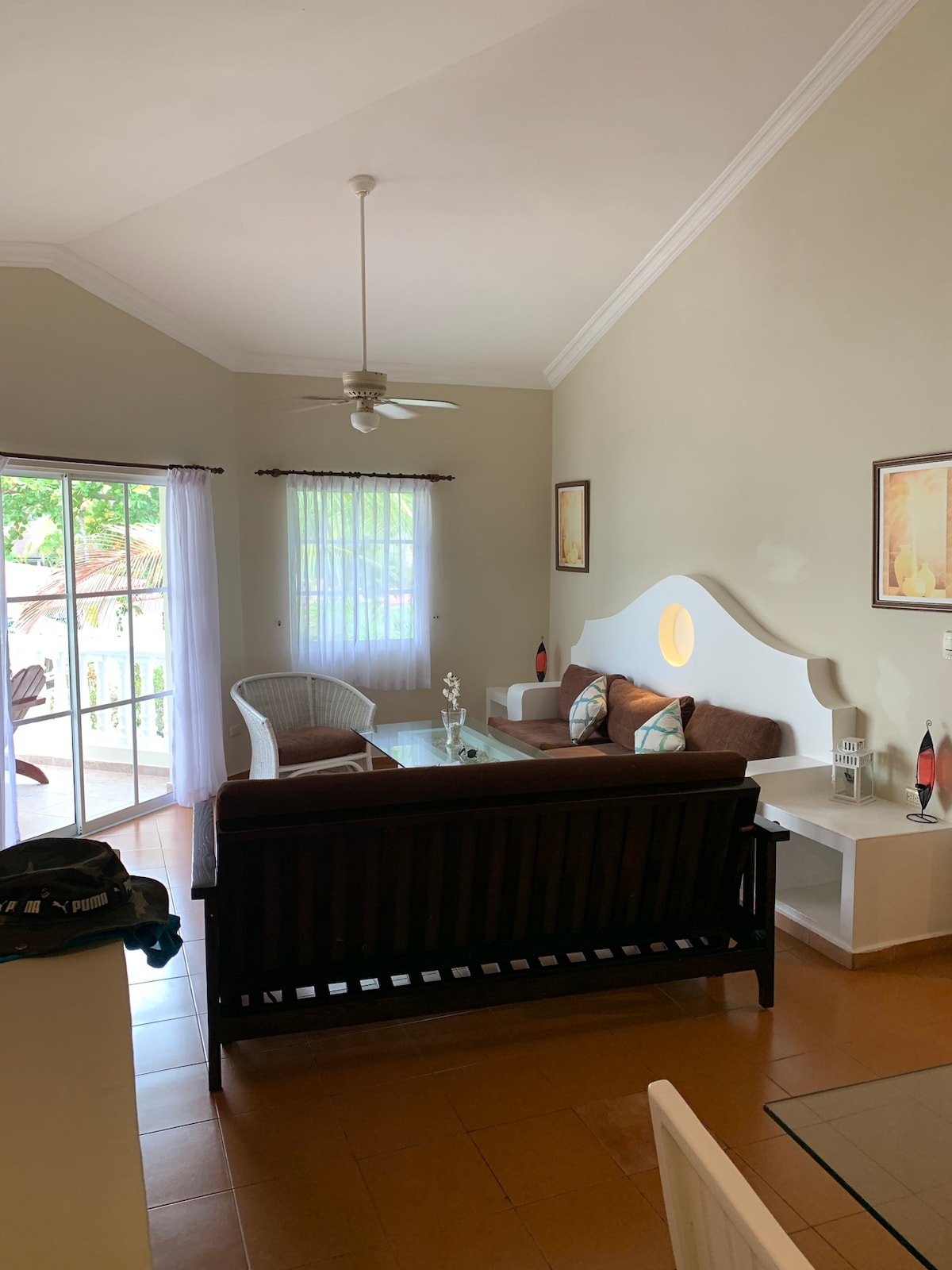 A spacious living area features a high ceiling with a ceiling fan and large glass doors leading to a balcony. Comfortable seating arrangements include a dark wooden sofa and light-colored chairs. Decorative wall art adds visual interest, and natural light filters through the window.