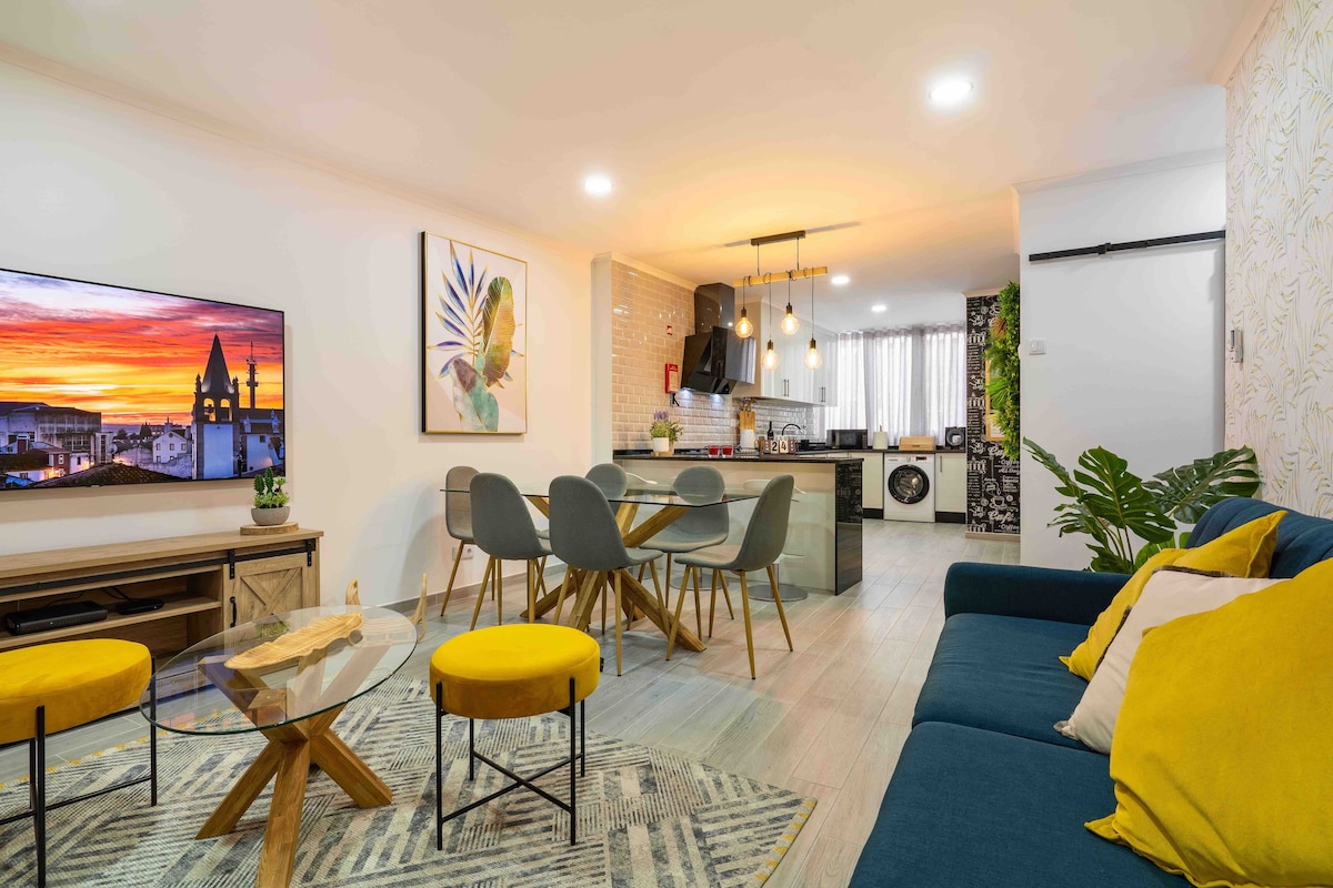 A modern living area features a blue sofa and a geometric area rug. A circular glass coffee table rests on the rug, surrounded by bright yellow chairs. A wall-mounted television displays a sunset view, while a dining area is visible in the background alongside the kitchen.