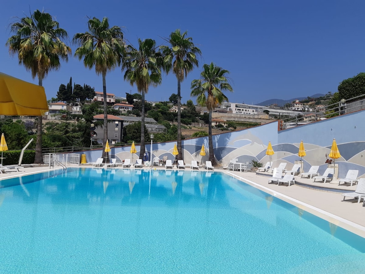 A large swimming pool is surrounded by palm trees, with numerous sun loungers positioned along its edge. Bright yellow umbrellas provide shade, and the inviting water reflects the clear blue sky.
