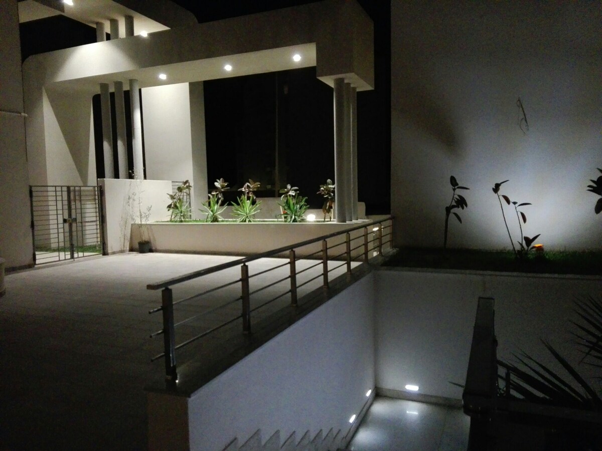 An exterior view of the building at night is shown, featuring modern architectural design elements. Soft lighting highlights the landscaped area with plants alongside a staircase. A railing provides safety along the walkway leading to the entrance, creating a serene and inviting atmosphere.