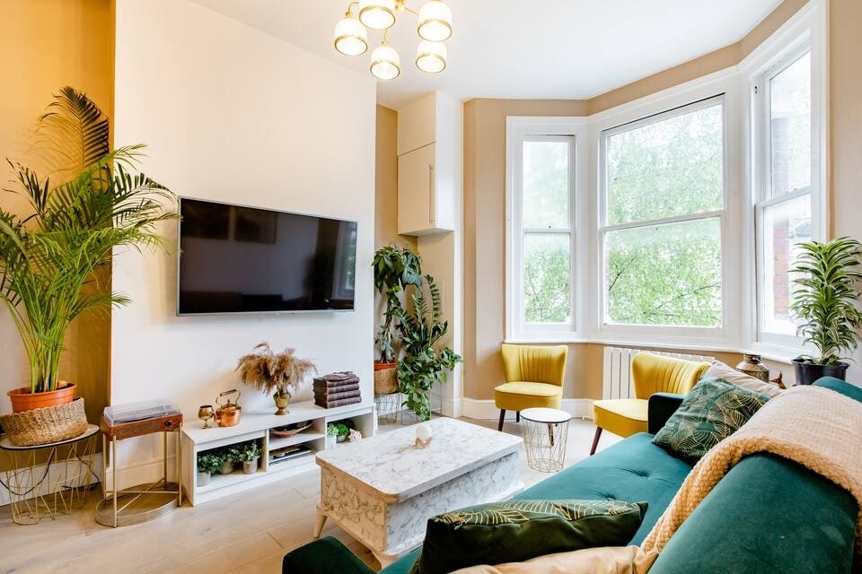 A living room is showcased with large bay windows allowing natural light. A modern TV is mounted on a light-colored wall. Green plants are positioned around the space, with two yellow chairs and a teal sofa enhancing the design. A coffee table is centered in front.