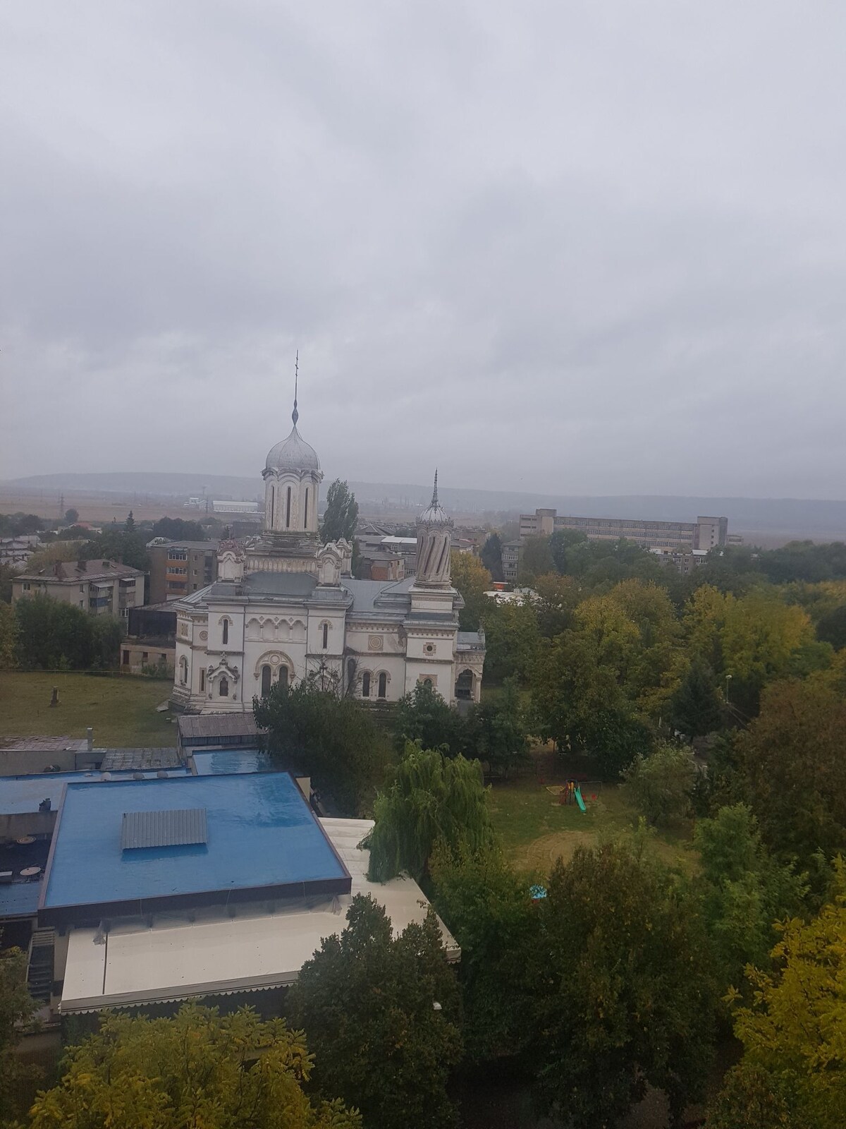 A cloudy sky looms over a historic building featuring intricate architectural details, including domes and spires. Surrounding lush trees and green spaces are visible, with modern structures in the background. A glimpse of a blue roof can be seen in the foreground, enhancing the scene's layers.
