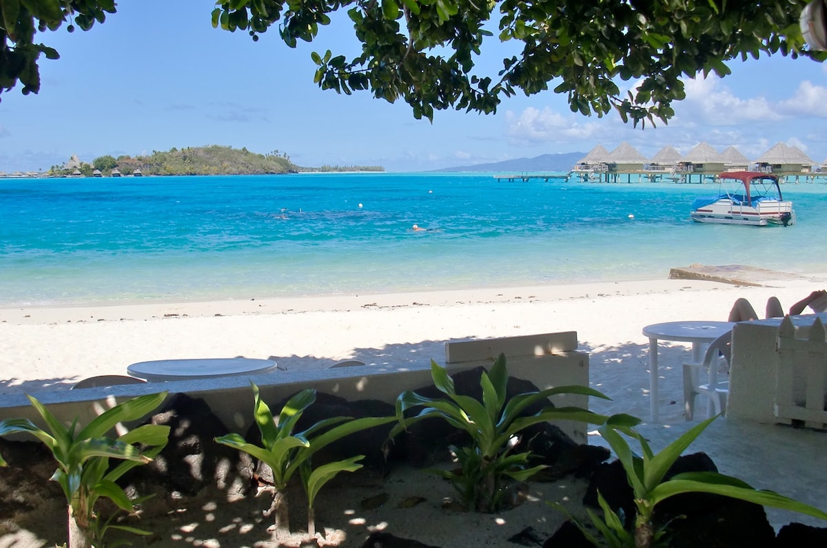 A serene beach view reveals soft white sand meeting clear turquoise water. Lush green foliage frames the scene, while distant overwater bungalows and boats on the water add depth. Gentle waves lap at the shore, creating a calm and inviting atmosphere.