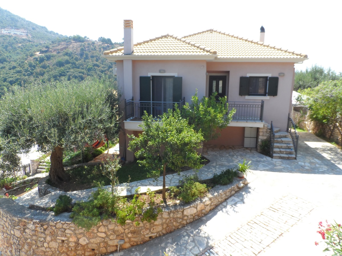 A charming house is nestled on a small hill, framed by lush greenery and gentle stone pathways. The exterior features a warm color palette, complemented by dark green shutters and a welcoming porch. Olive trees and carefully placed plants enhance the tranquil setting.