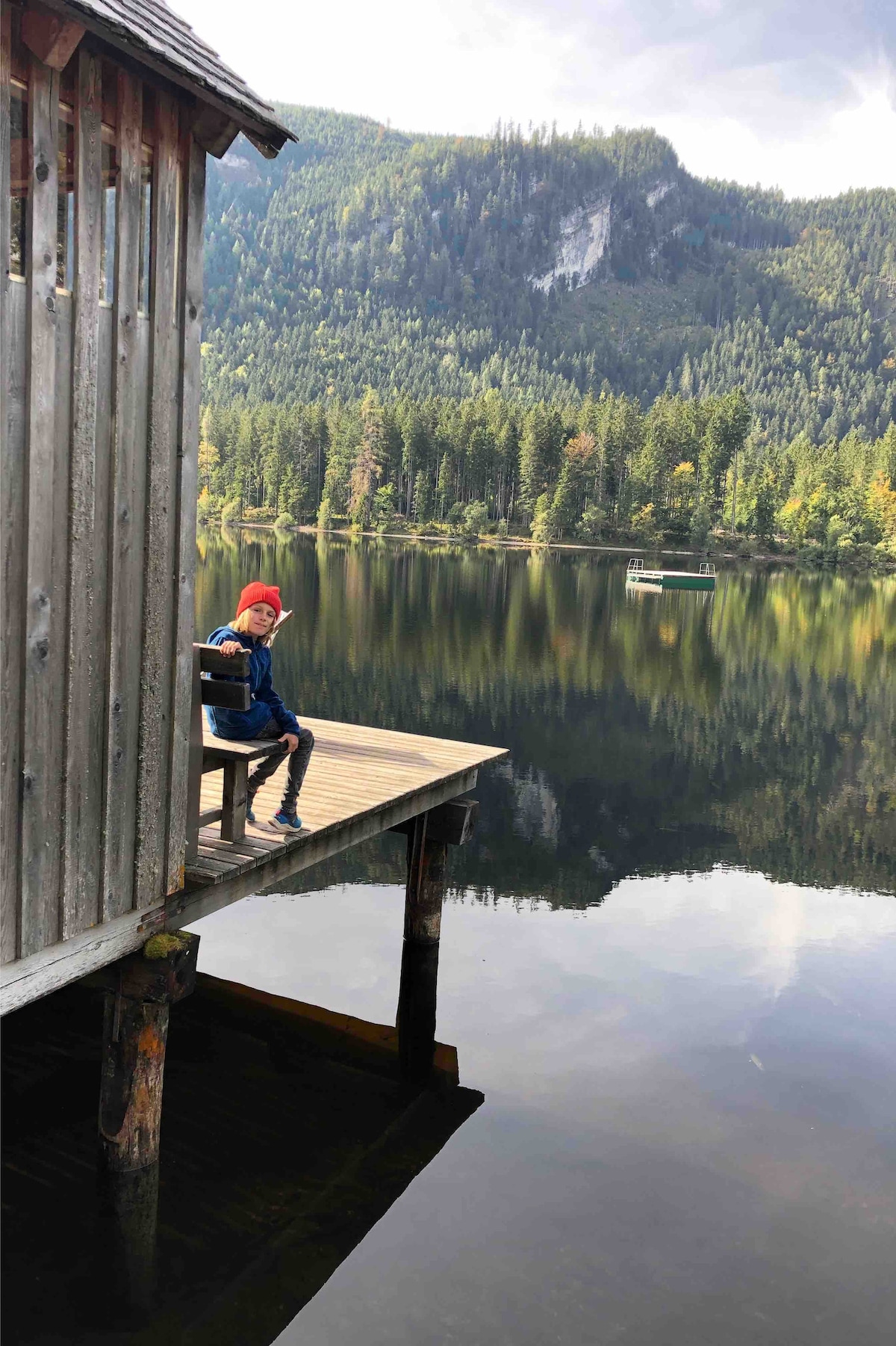 A wooden dock extends over a calm lake, reflecting surrounding mountains and lush evergreen trees. A figure sits on a bench at the dock's edge, gazing towards the serene water, creating a peaceful and harmonious scene.