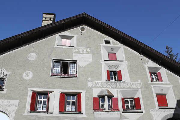 The façade of a historic Engadin house is displayed, featuring decorative wall motifs and red wooden shutters. Multiple windows are visible, adding symmetry to the building. The clear blue sky serves as a backdrop, highlighting the architectural details of this traditional multi-family residence.
