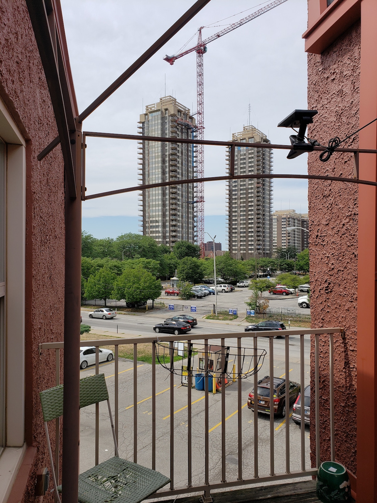 The outdoor seating area features a small balcony with two green chairs and a clothes drying rack. A view of the cityscape is visible, showcasing two tall buildings under construction, with trees and parking areas in the foreground.