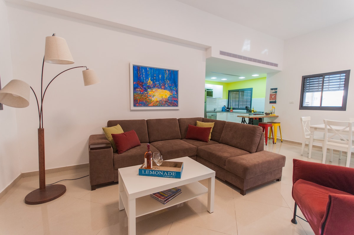 A spacious living area is furnished with a sectional sofa featuring vibrant cushions. A coffee table sits in the center, accompanied by a decorative piece. A colorful artwork adorns the wall, while the kitchen and dining area are visible in the background.