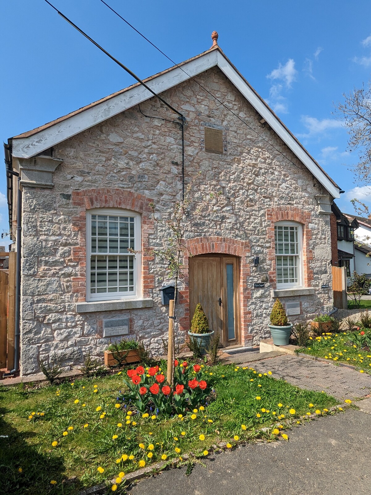 The exterior of a converted chapel features a stone facade with red brick accents. Two windows are symmetrically placed on either side of a wooden door, surrounded by flowerbeds of tulips and greenery. A clear blue sky is visible above, adding to the tranquil setting.