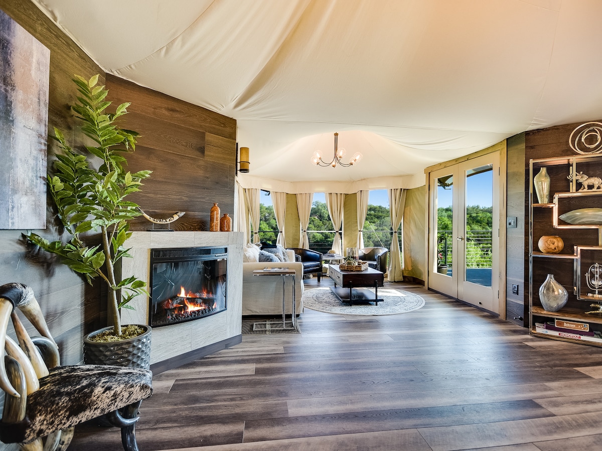 A spacious living area features a cozy electric fireplace against a wooden wall, offering warmth. Large windows are adorned with flowing drapes, allowing natural light to fill the room, while comfortable seating arrangements invite relaxation. Contemporary decor elements are arranged thoughtfully throughout the space.