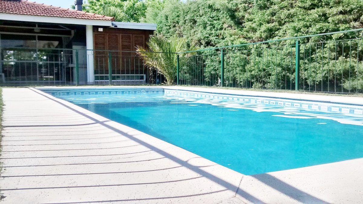 A private swimming pool is surrounded by a safety fence, offering a clear view over the water. The pool area features well-maintained paving, with trees providing greenery in the background. Sunlight reflects off the water, creating a calming atmosphere.